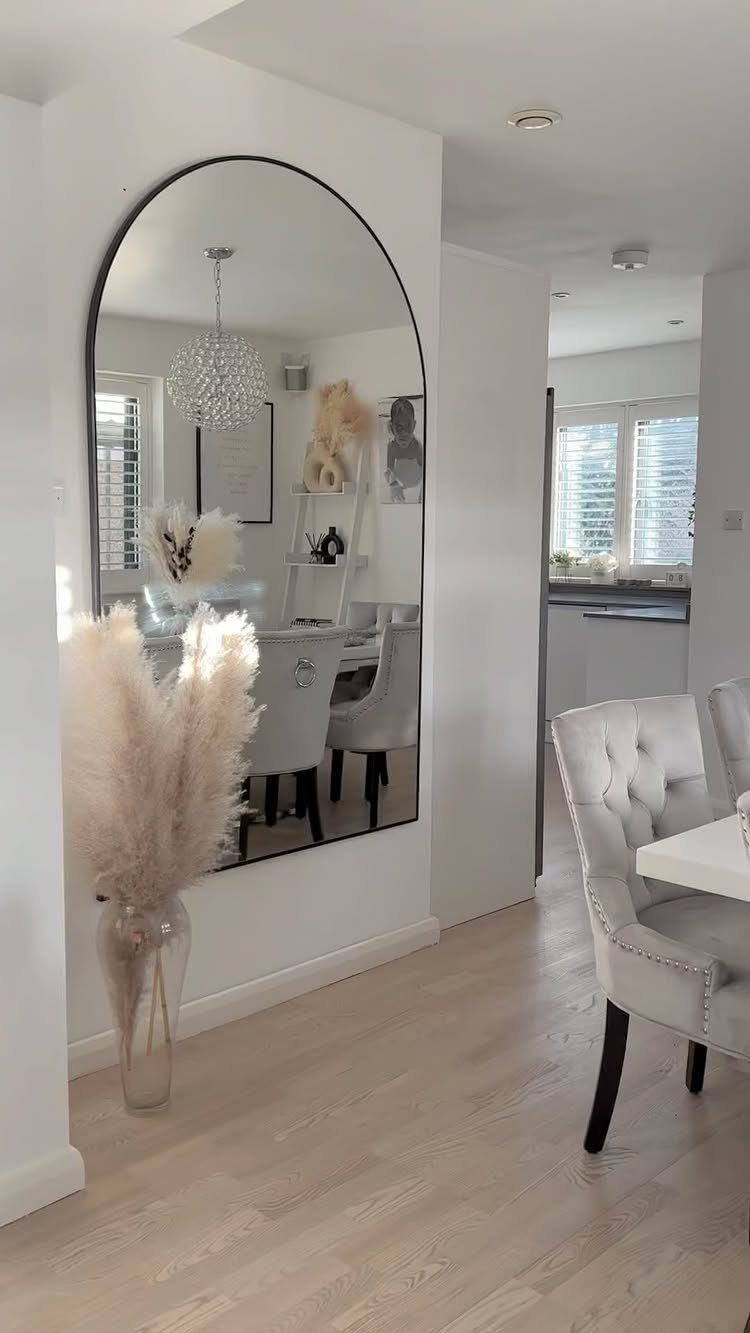Arched wall mirror reflecting a bright dining room with white tufted chairs.