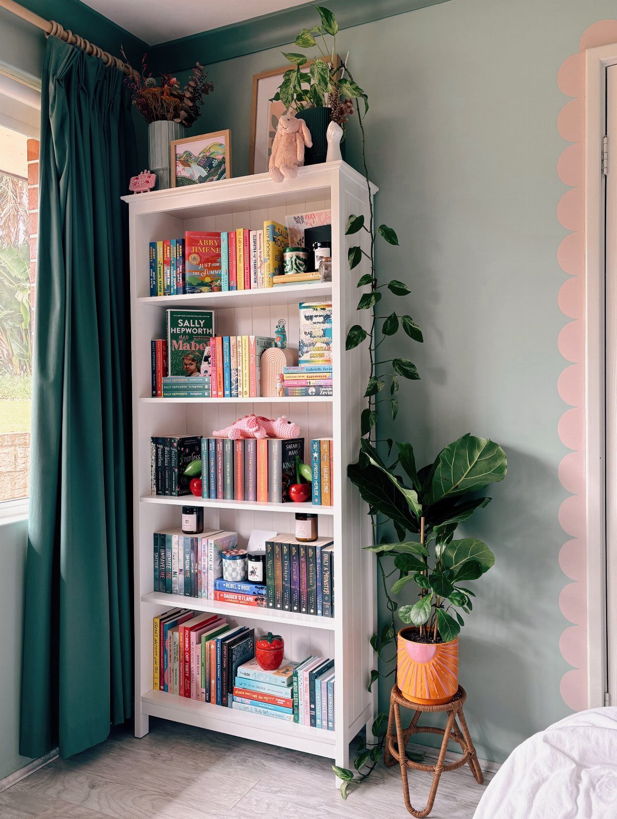 White bookcase filled with colorful books and plants against a light green wall.