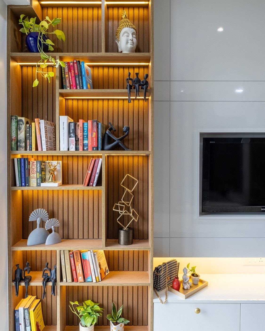 Light-wood wall-mounted bookcase with integrated lighting, books, and decorative sculptures in a living room.