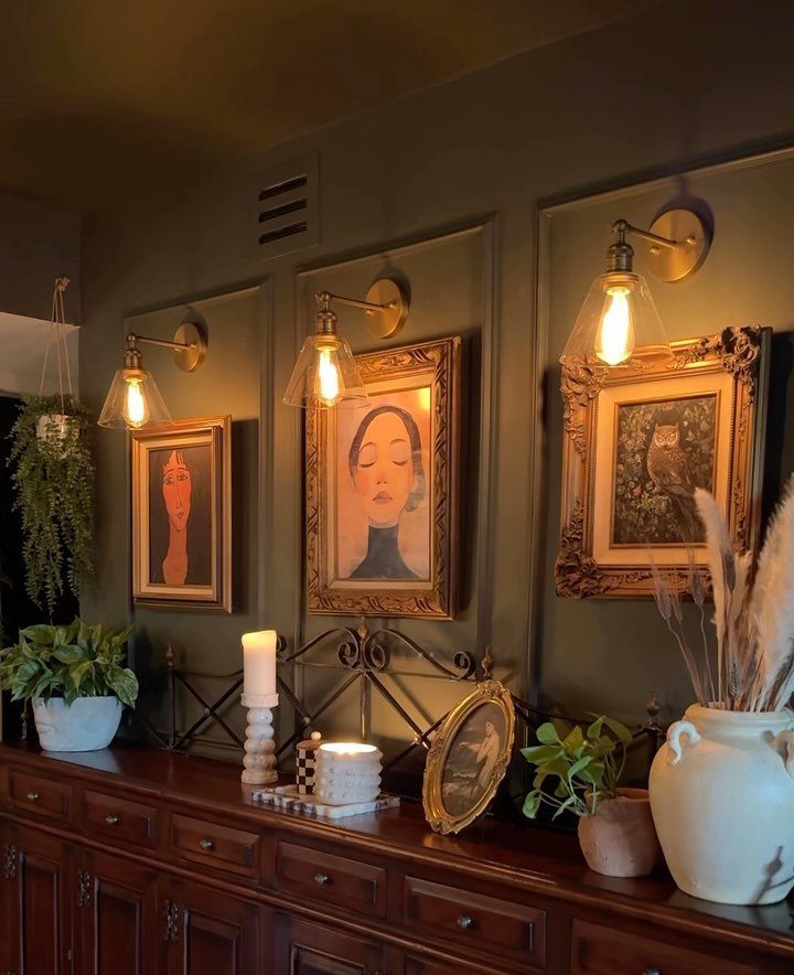 Dark green wall with gold-framed art, brass sconces, and a dark wood sideboard.