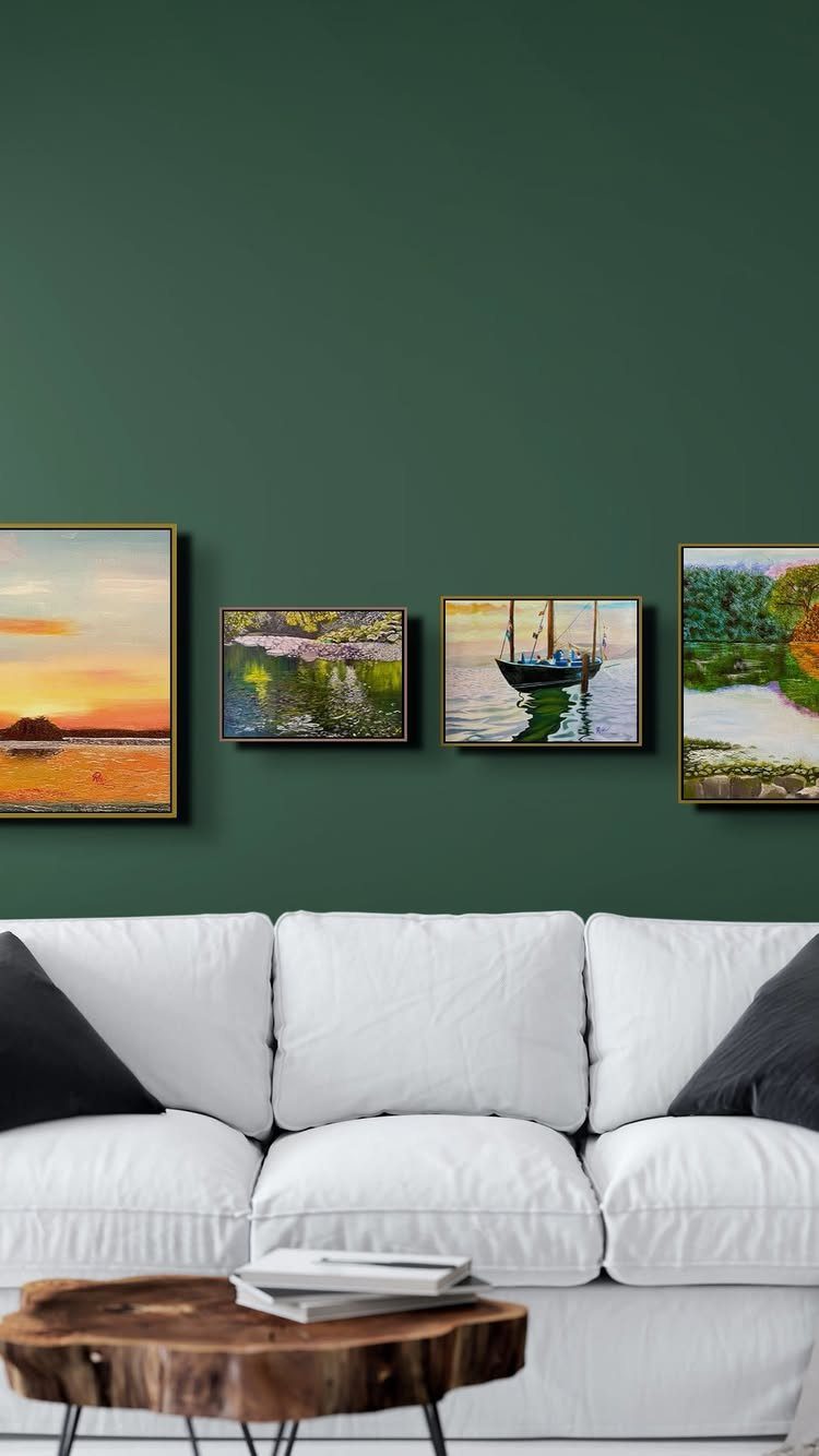 Four framed landscape paintings on a dark green wall above a white sofa.