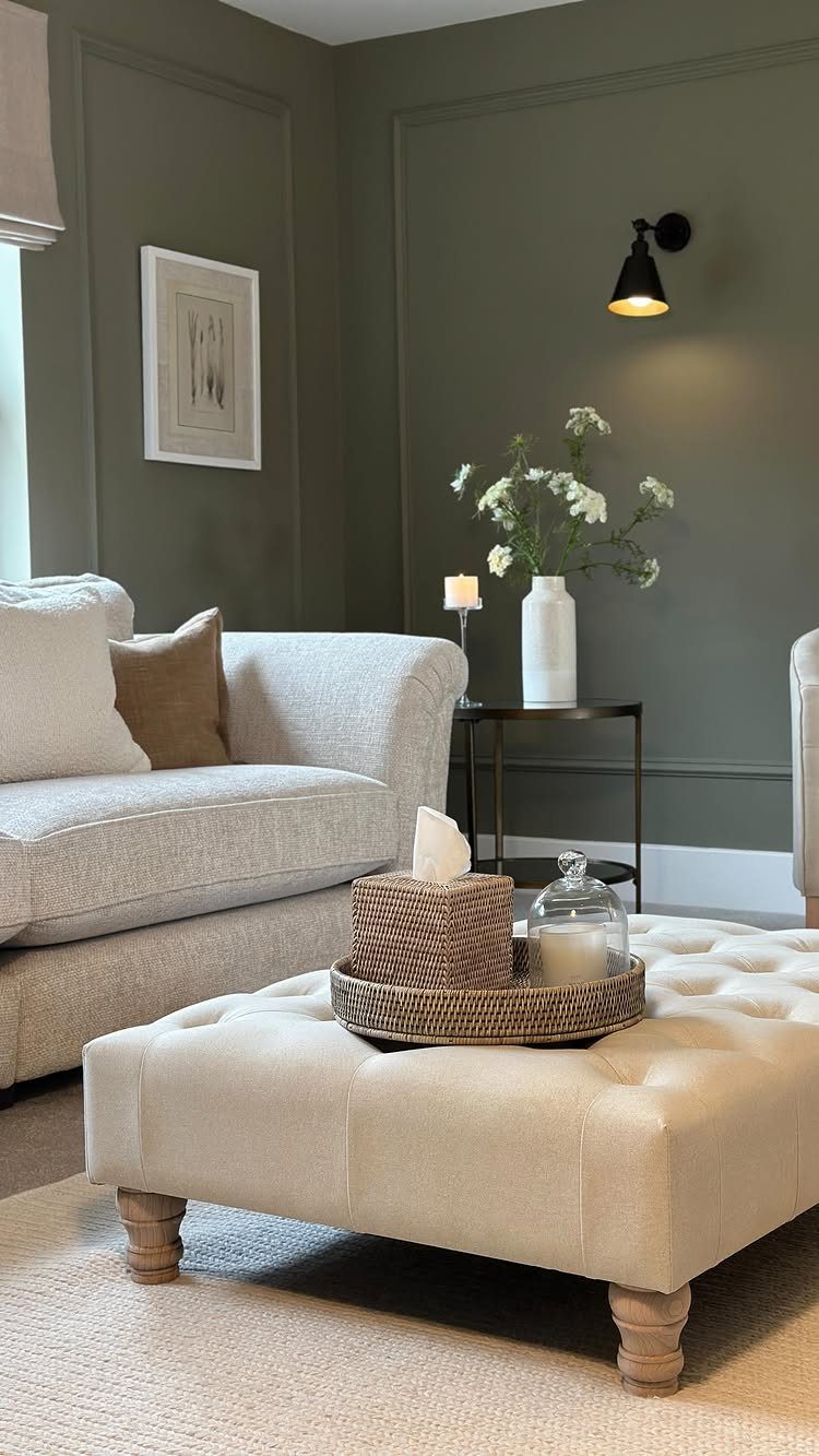 Dark olive green living room with beige sofa, tufted ottoman, and wall sconce.