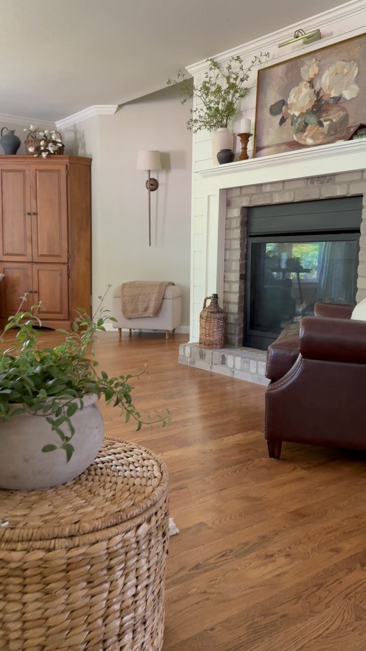 Farmhouse living room with leather chair, wicker ottoman, and brick fireplace hearth.
