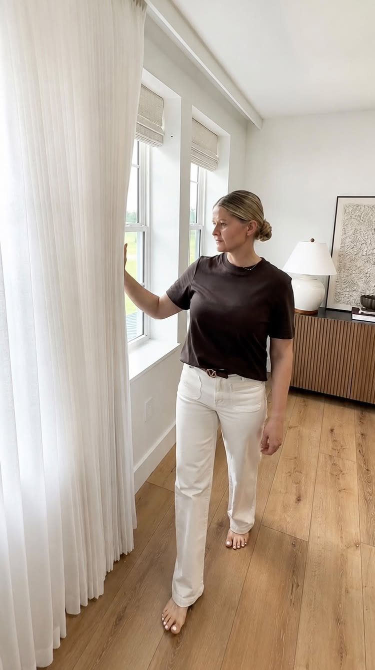 Woman touching long white sheer linen curtains in a bright living room.