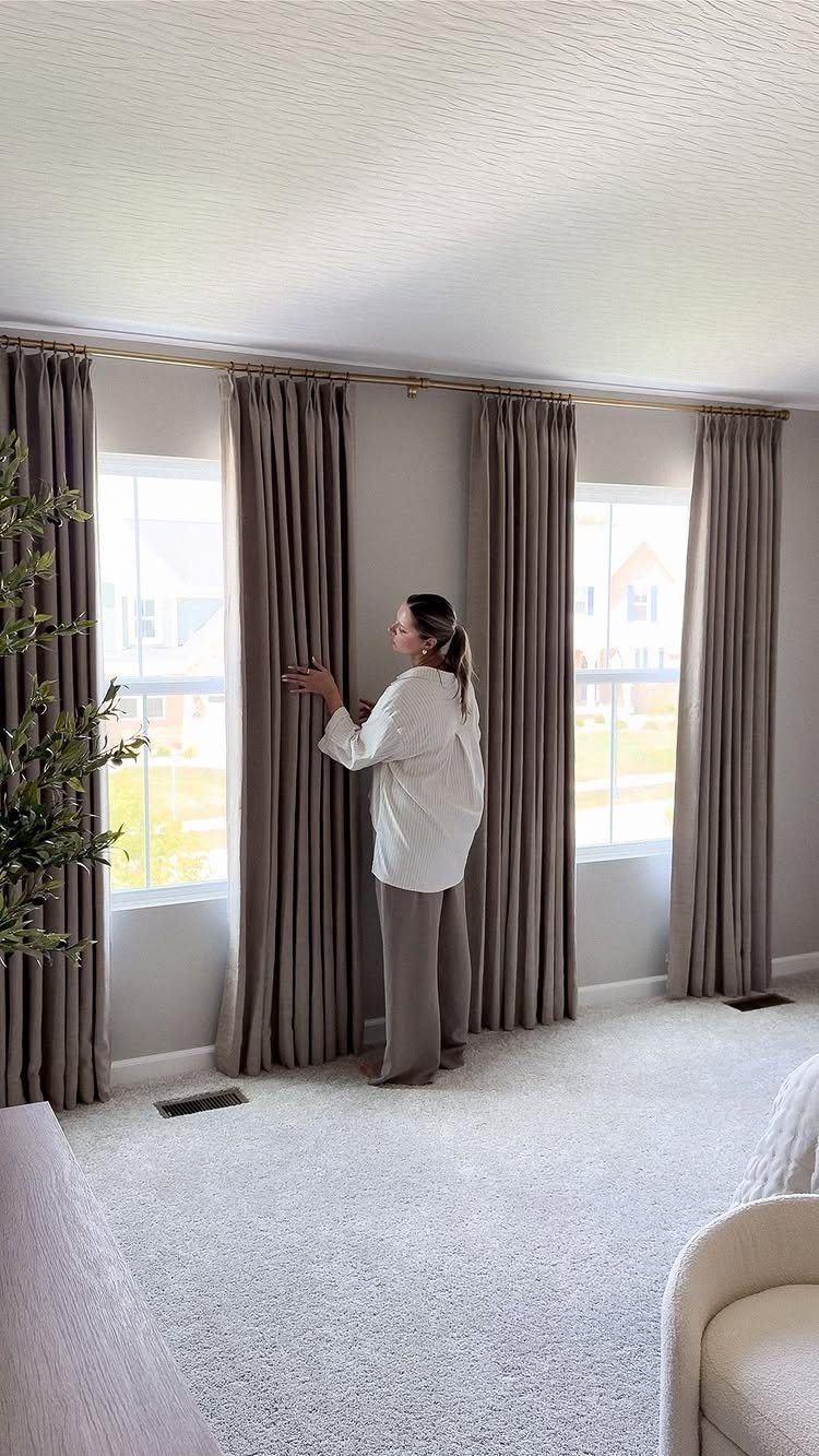 Woman adjusting taupe curtains that extend beyond the window frame in a living room.