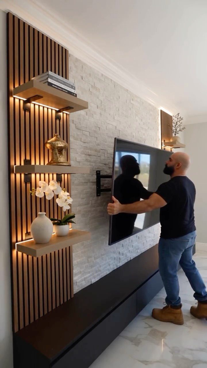 Man installing a TV on a white stone wall with wood slat shelving.
