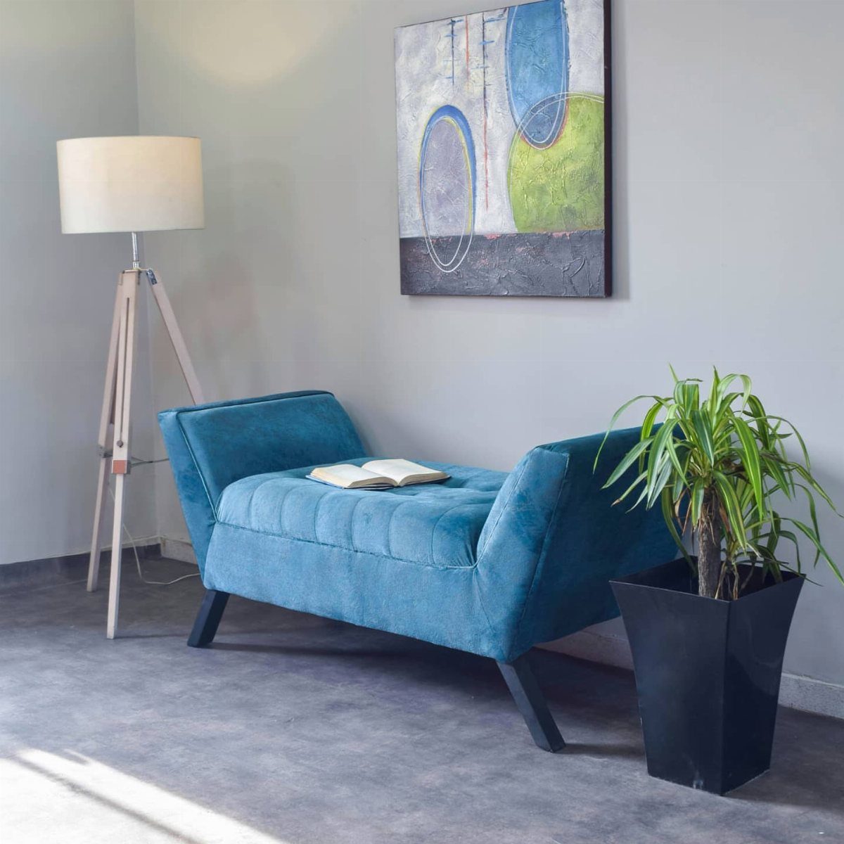 Blue velvet chaise lounge with a book, lamp, and plant in a living room.