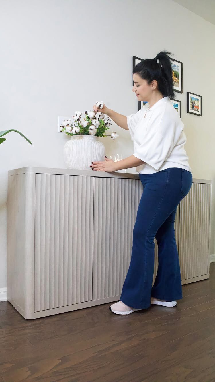 Woman arranging white flowers on a light grey ribbed sideboard in a living room.