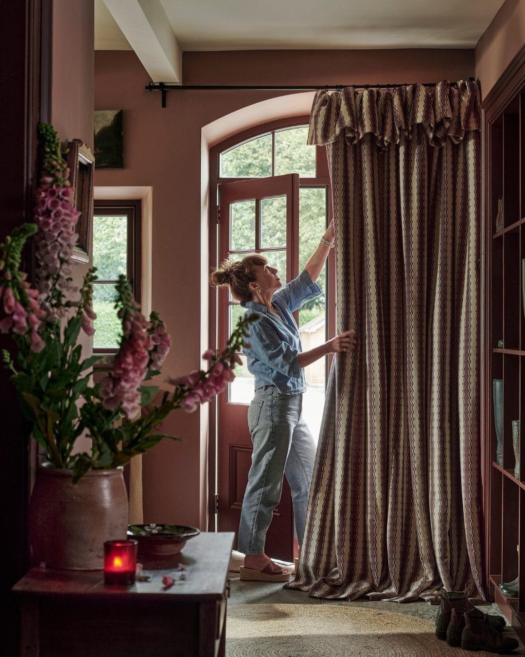 Woman adjusting brown patterned curtains with a ruffled flop-over frill valance in a room.