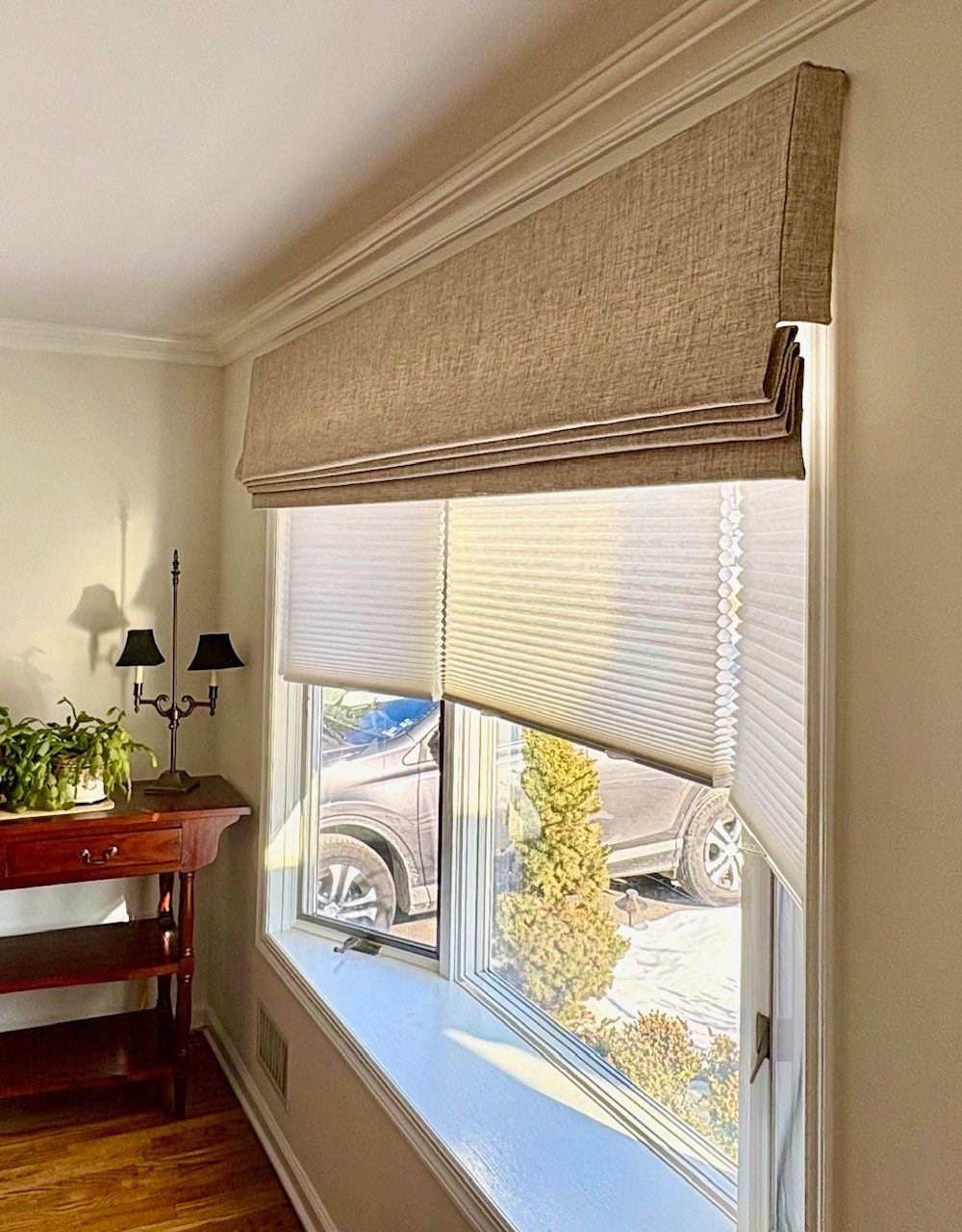 Beige textured Roman shade valance hanging over white cellular blinds in a bright room.