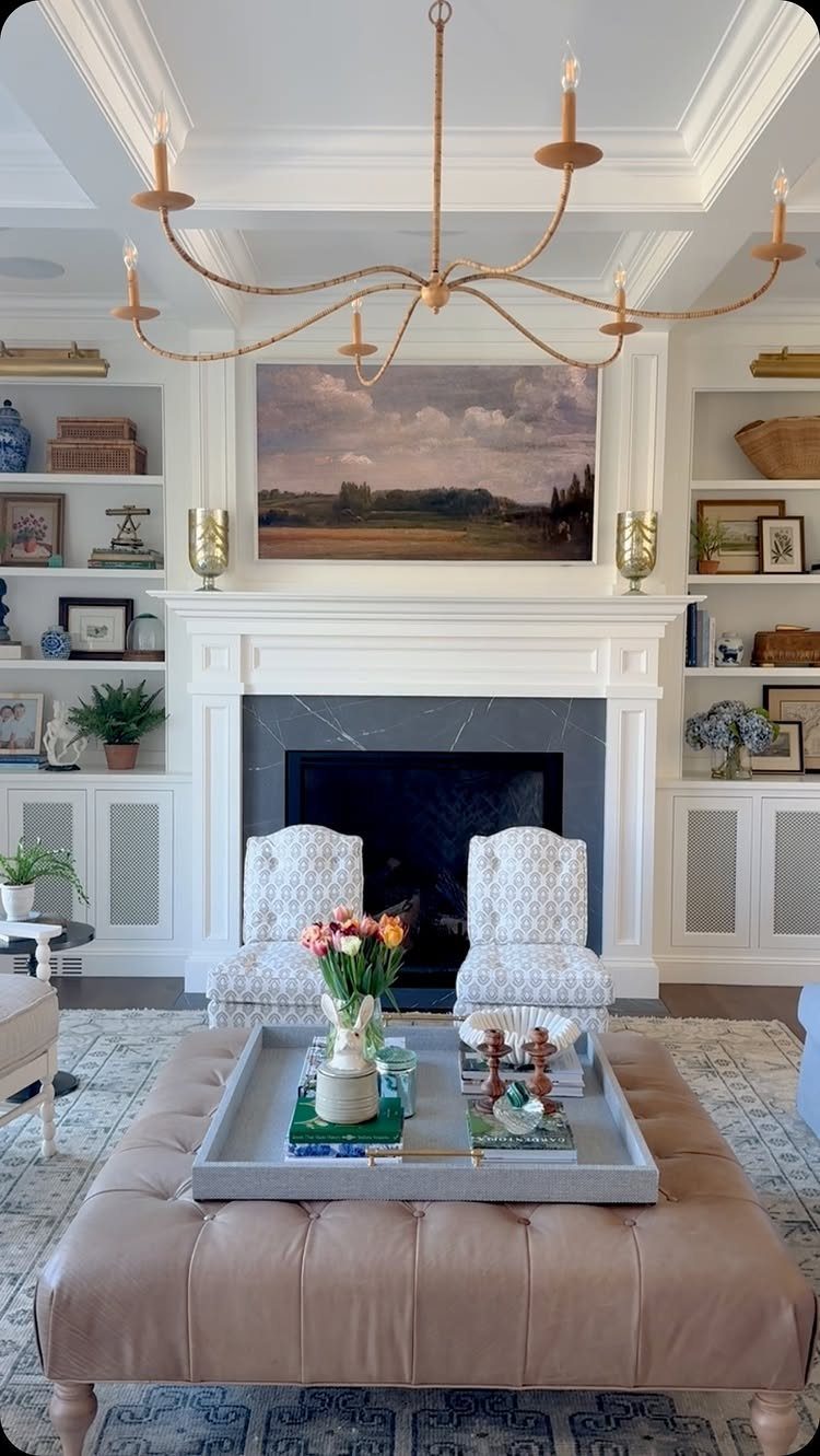 White fireplace with built-in bookshelves, landscape art, and tan leather ottoman in living room.