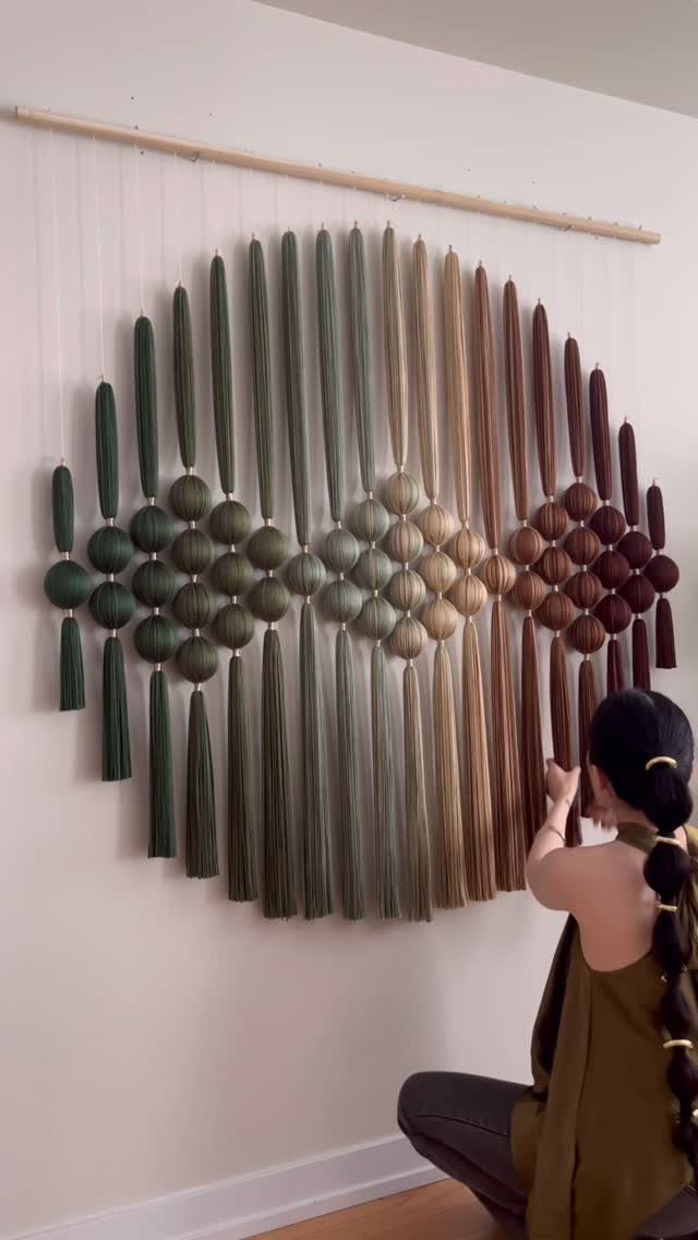 Textile wall hanging with green and brown yarn tassels and spheres on a white wall.