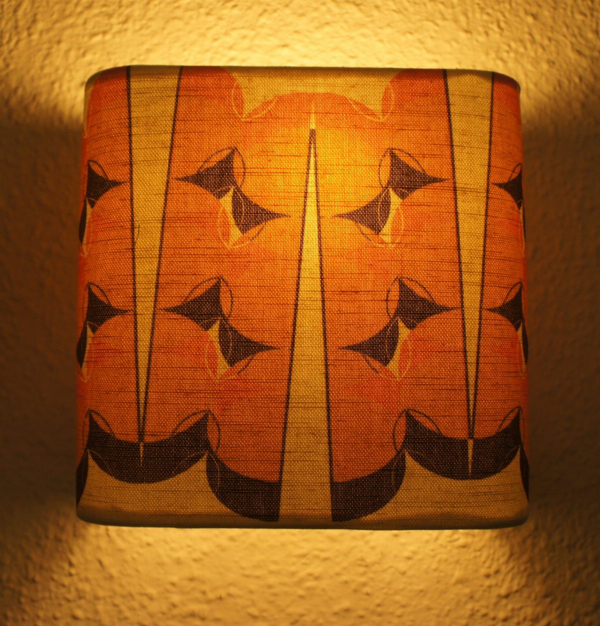 Lit wall lamp with an orange patterned fabric shade against a beige wall.